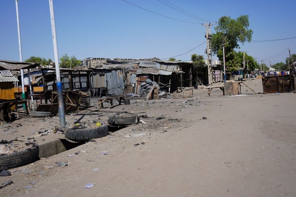 Dusty street with scattered debris, abandoned market stalls, and a few trees under a clear blue sky.