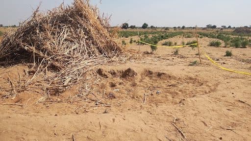A mound of dry brush next to a fenced-off sandy area in an open field under a clear sky.