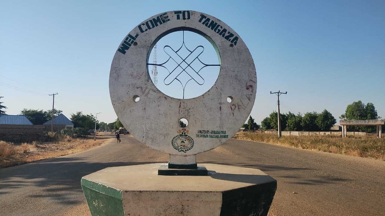 Welcome sign in Tangaza, Nigeria, with a circular design in the center. Road, trees, and buildings are visible in the background.