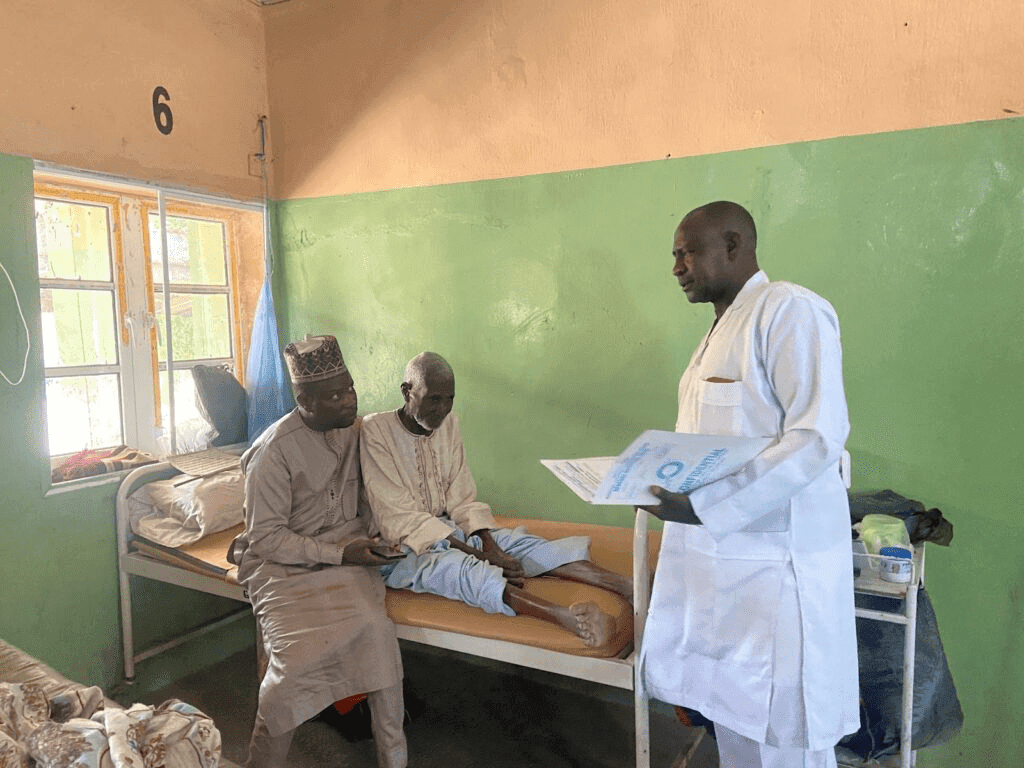 A doctor in a white coat talks to two men, one seated on a hospital bed, in a room with green walls.
