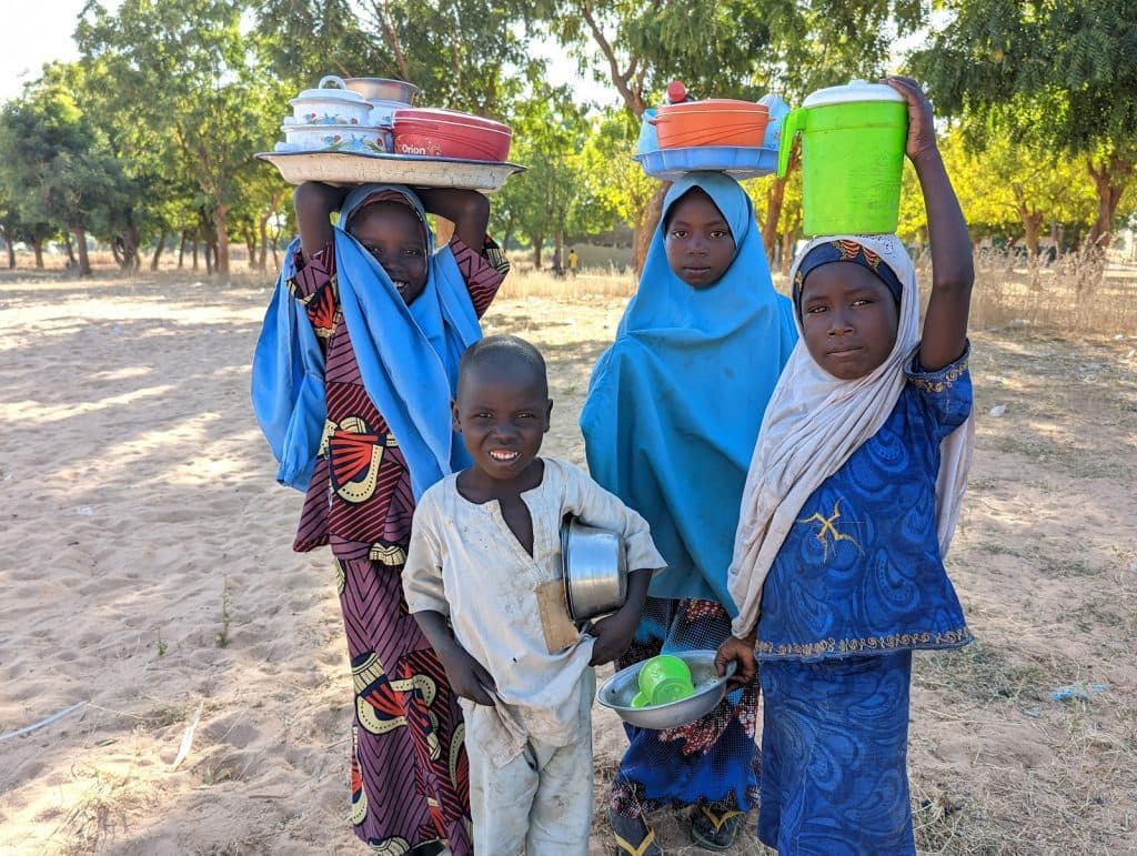 Four children outdoors, three wearing colorful clothing and carrying items on their heads, one smiling with a pot. Trees in the background.