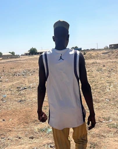 Man in a white sleeveless shirt and striped pants walks on a dry field under a clear blue sky, with buildings in the background.