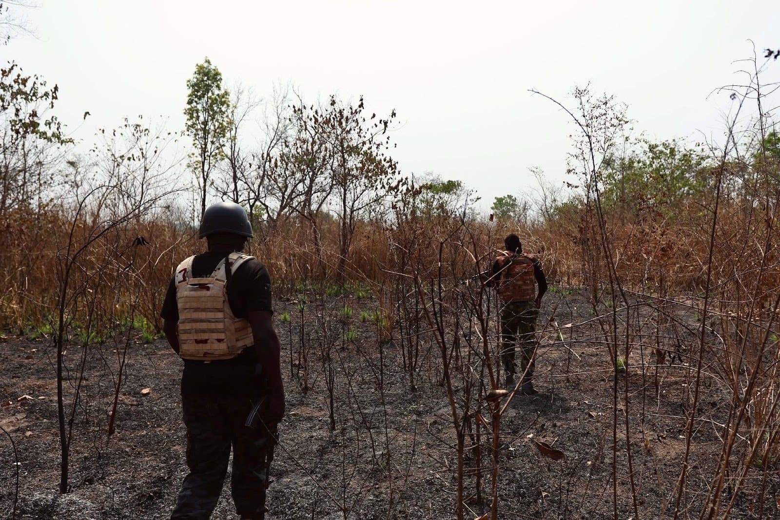 Two soldiers in protective gear walk through a dry, wooded area with sparse vegetation under a clear sky.
