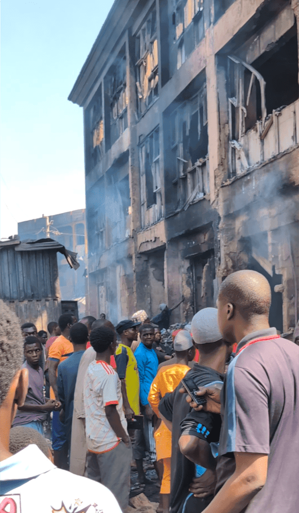 Crowd gathers at a damaged building with smoke, assessing fire aftermath.
