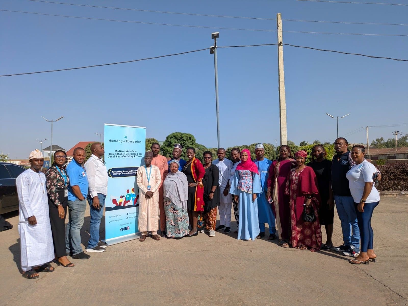 A group of people standing together outdoors near a banner for a multi-stakeholder roundtable on local peacebuilding efforts.