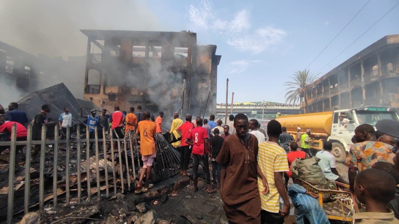 Crowd gathers around a smoke-filled, fire-damaged building as a tanker arrives; debris lies scattered on the ground.