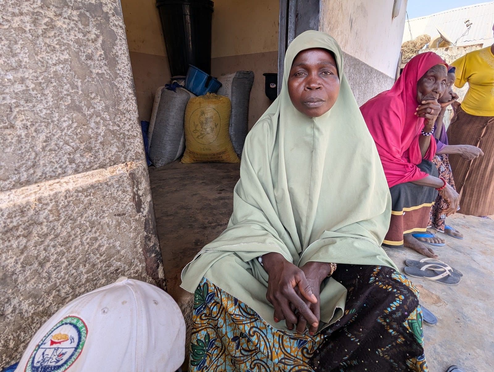 A woman in a green dress and headscarf sits outside a building, with colorful sacks of grain behind and two women sitting nearby.