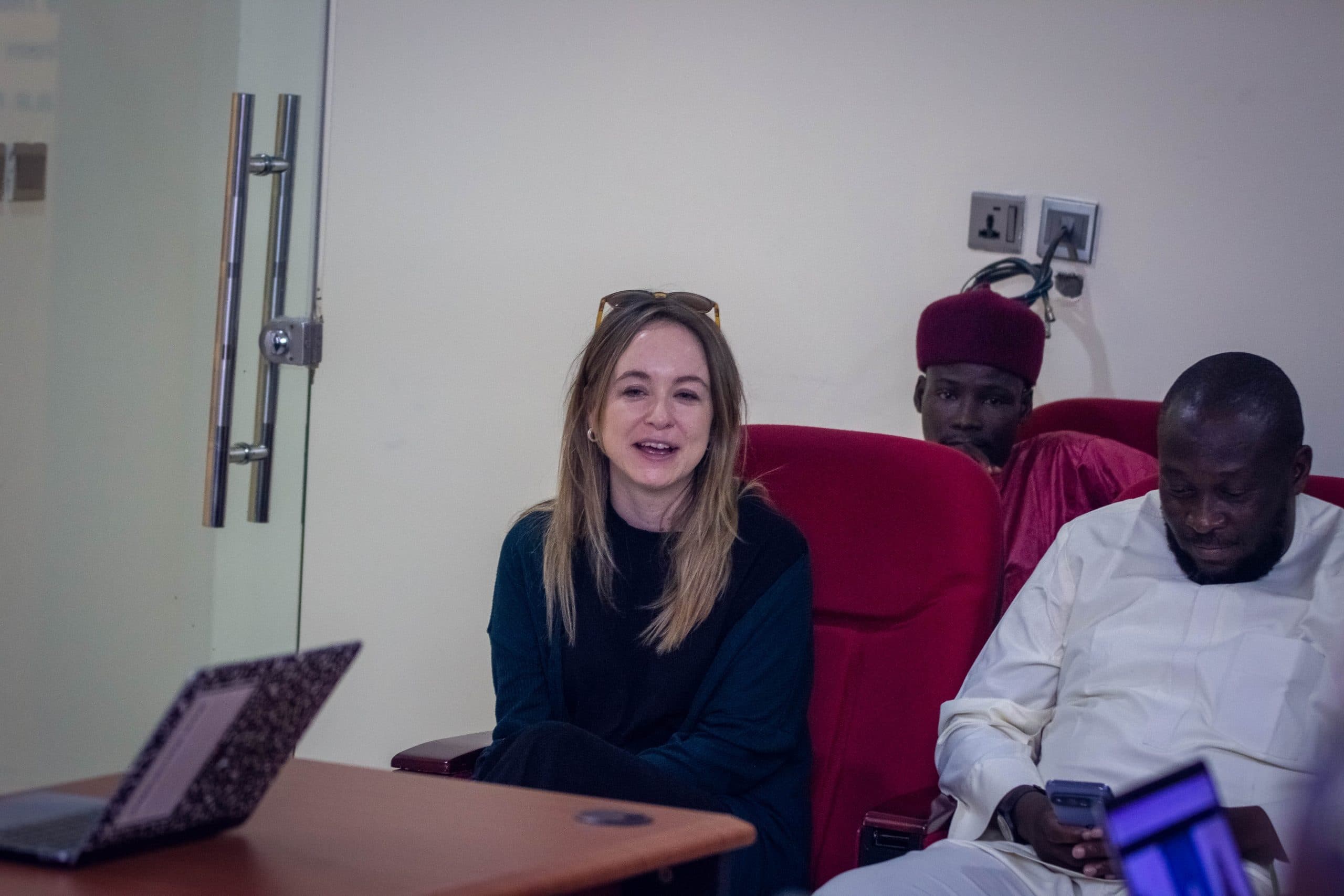 Three people seated, one woman smiling, two men beside her, one using a phone. A laptop is open on the table.