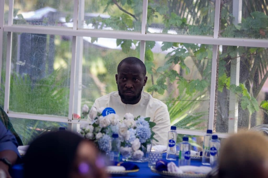 Man seated at a table with white flowers and bottled drinks in the foreground, greenery visible through nearby window.