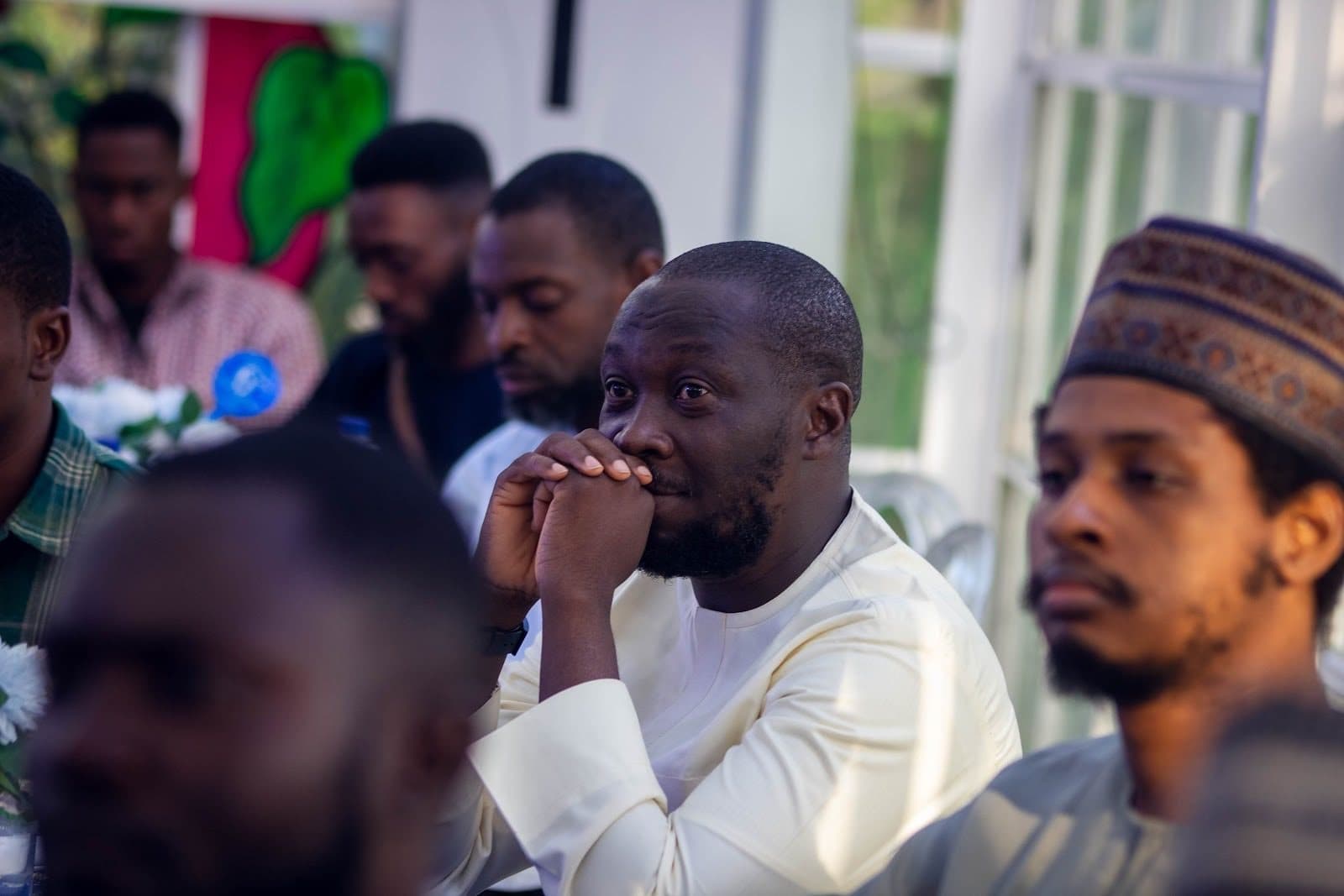People seated at a gathering, with one person attentively listening, hands clasped near their face.