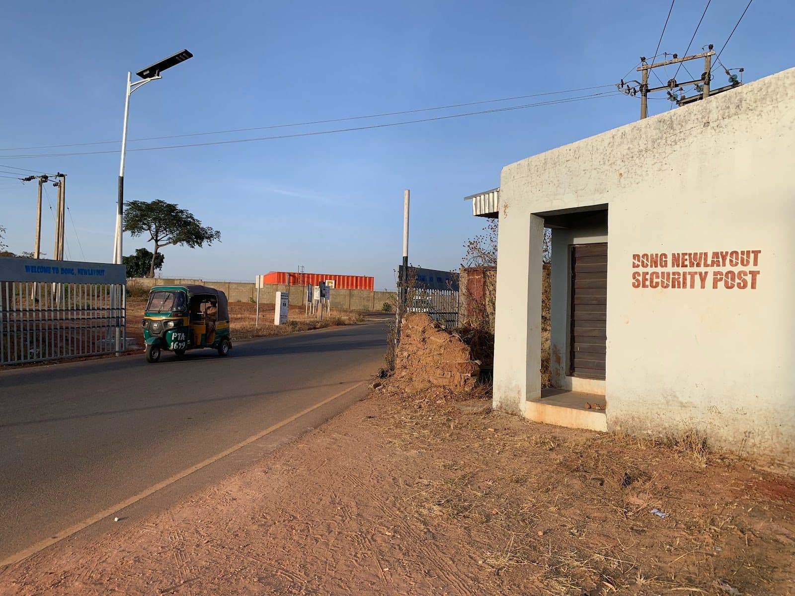 A tuk-tuk drives past a security post labeled "DONG NEW LAYOUT SECURITY POST" on a sunny day, with a gate in the background.