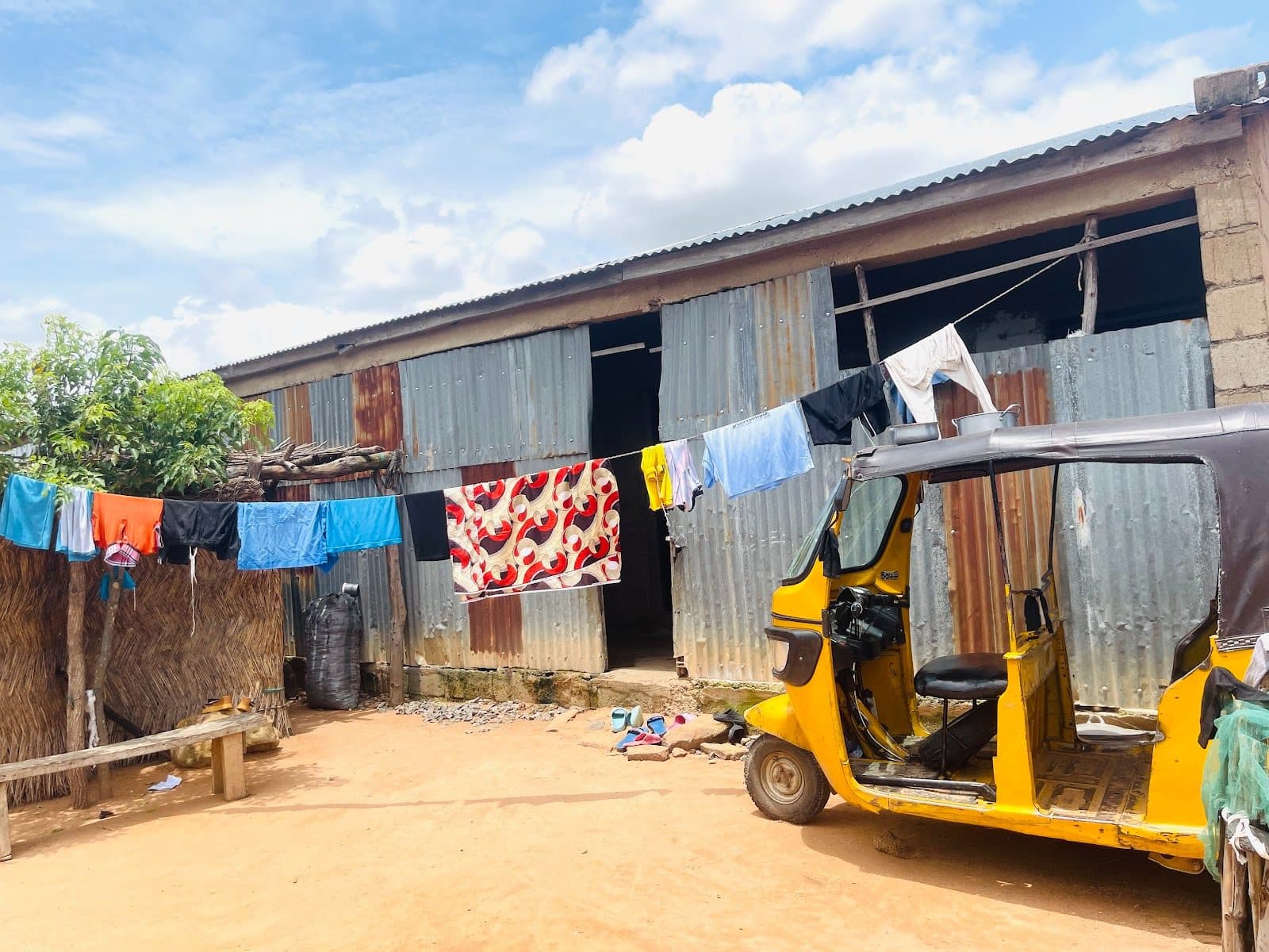 Yellow tuk-tuk parked outside a corrugated metal building, with colorful clothes hanging on a line, under a partly cloudy sky.