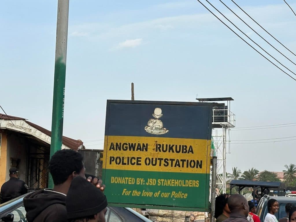 Sign for Angwan Rukuba Police Outstation with people nearby. Text mentions JSD Stakeholders
