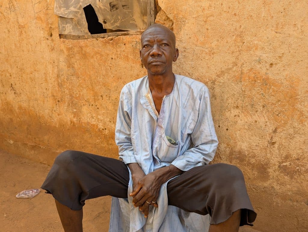 Man in a light blue shirt and dark shorts sits against a textured, weathered wall, looking straight ahead, with hands clasped.