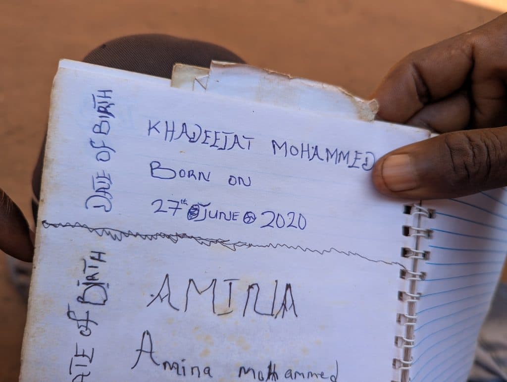 Close-up of a notebook page with handwritten text reading "Khadeejat Mohammed, born on 27th June 2020," held by a person