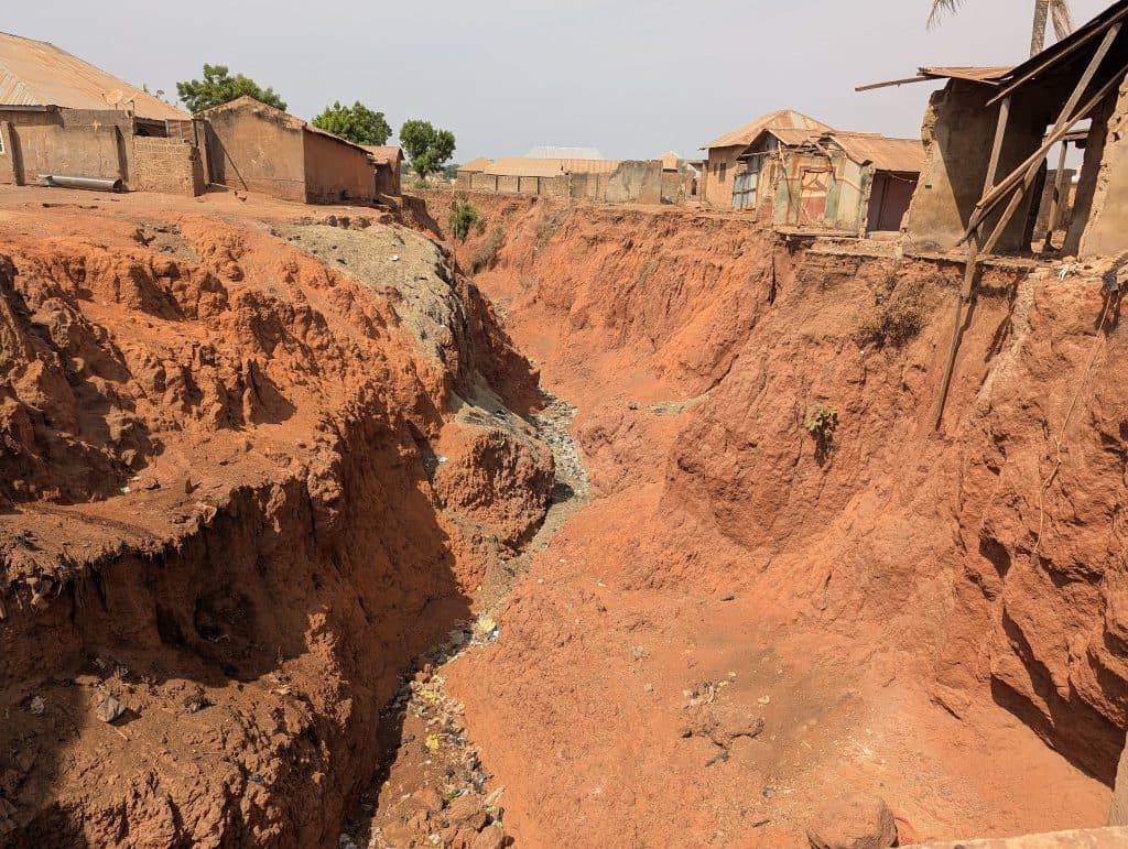 Erosion forms a deep gully between houses with rust-colored soil and debris scattered, highlighting the impact on nearby structures.