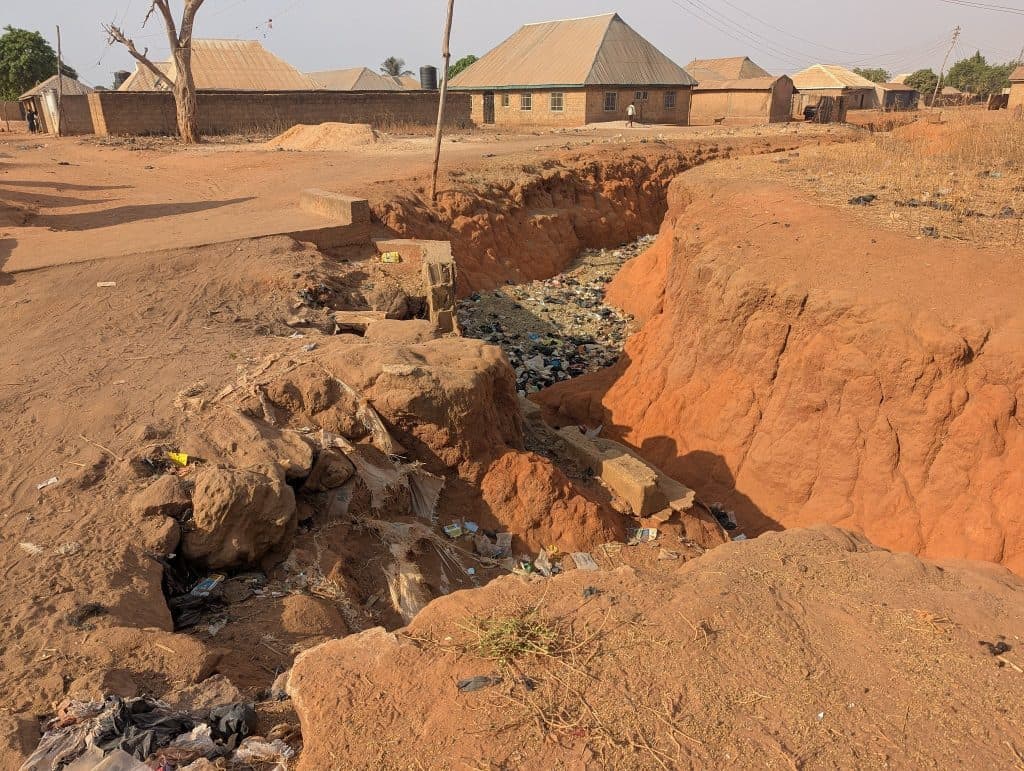 Erosion creating a deep gully with scattered trash in a dry, rural area near several small houses and buildings.