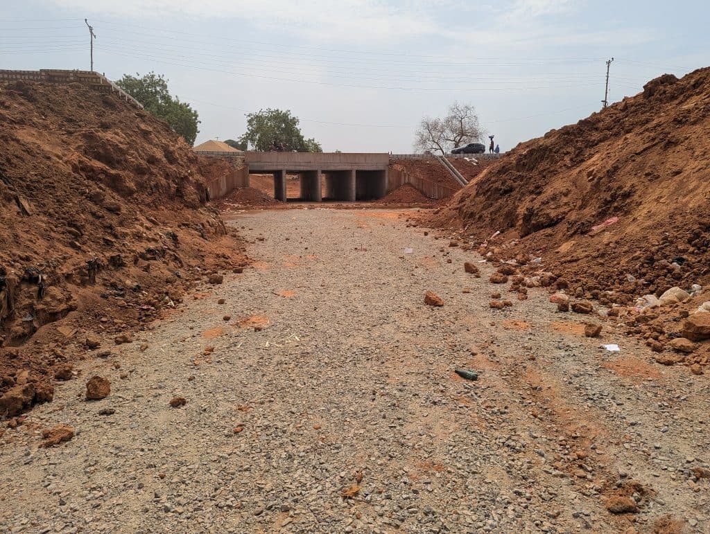 A dirt and gravel road leading to a concrete bridge, surrounded by piles of earth. Two people and a car can be seen on the bridge.