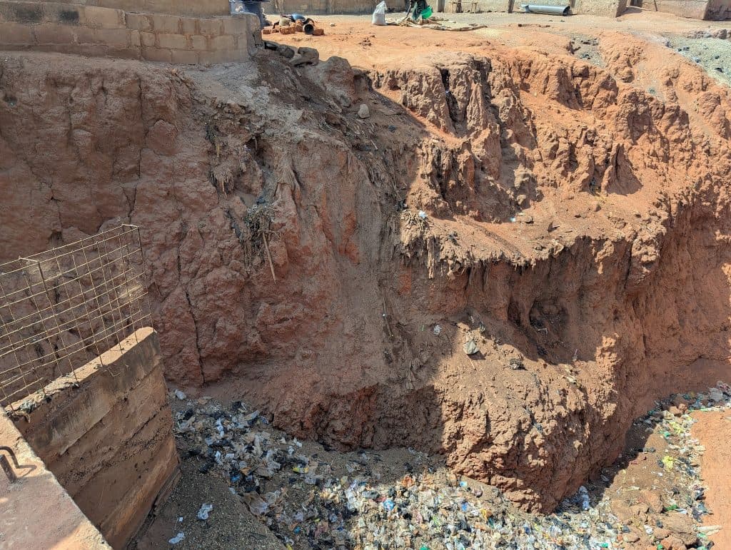 Eroded dirt cliff with strewn trash at base, next to a brick structure and metal fence.