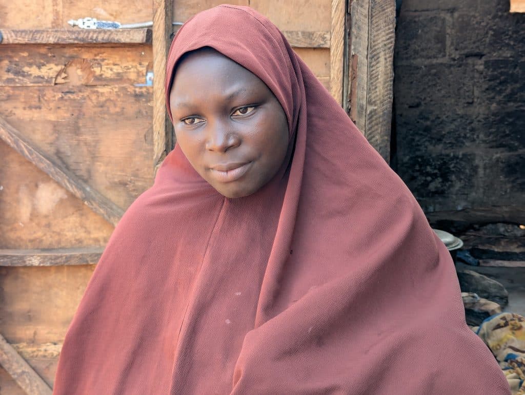 A person wearing a maroon headscarf sits in front of a wooden backdrop, looking thoughtful.