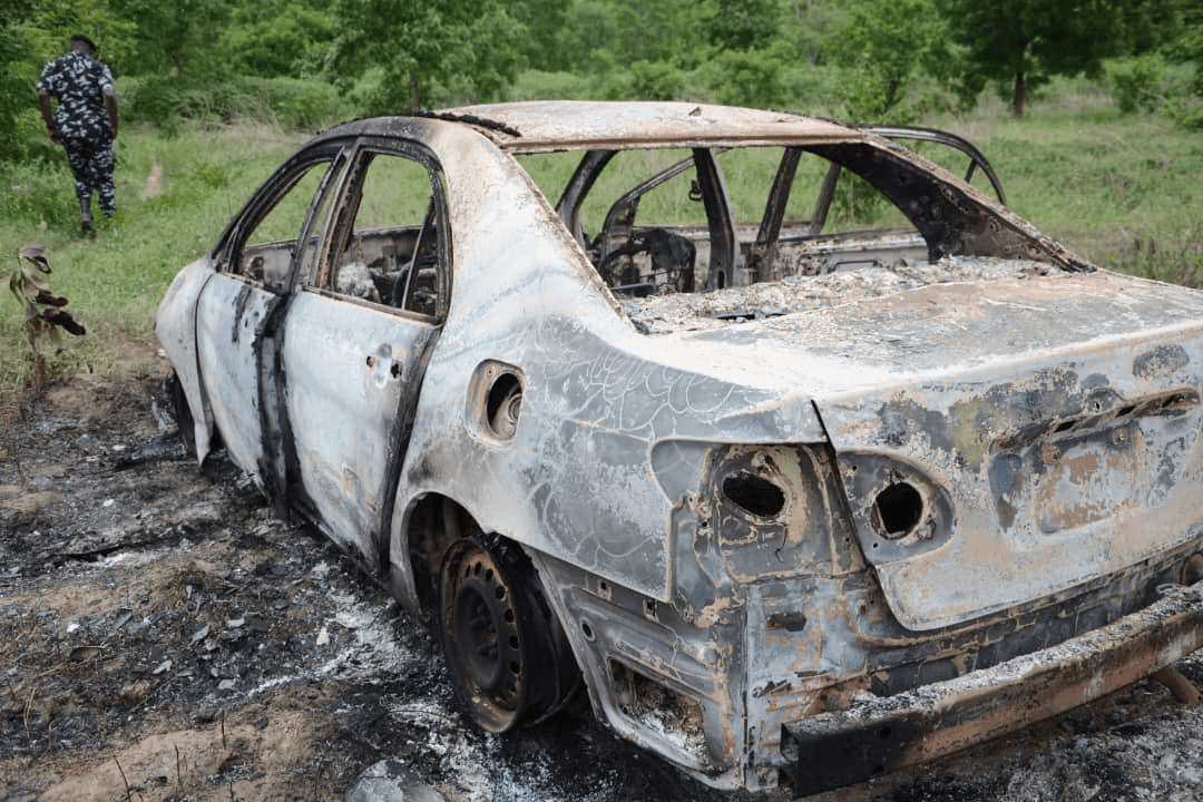 Charred remains of a burnt-out car in a grassy area with a person in patterned attire walking away in the background.