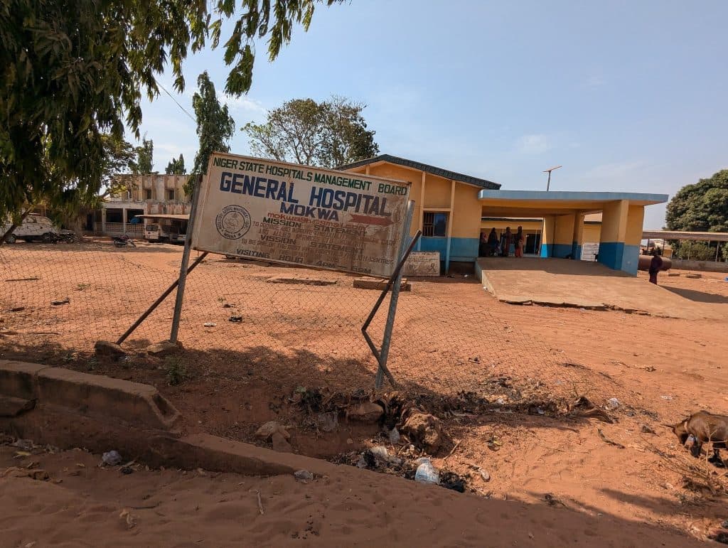 General Hospital Mokwa sign and entrance, with people gathered under a shade. Dry, sandy ground and trees in the background.