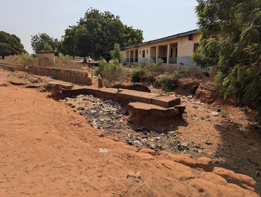 Dusty landscape with a bridge over a garbage-filled ditch, flanked by trees and a weathered building.