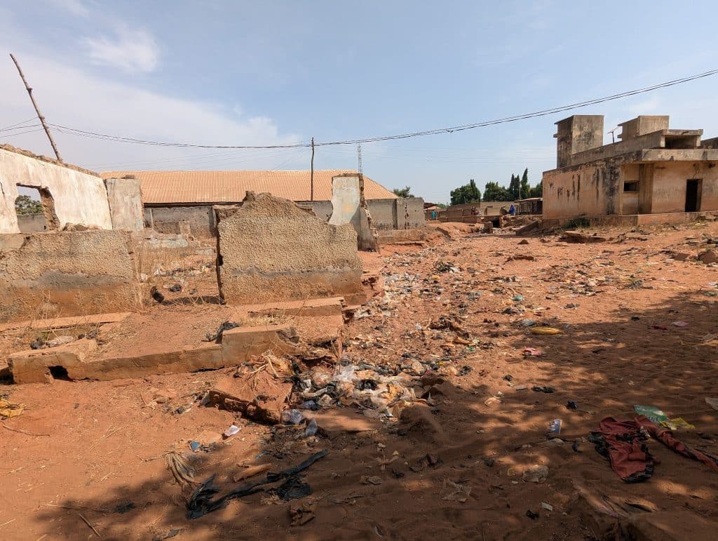 Dilapidated buildings in a dry, littered area under a partly cloudy sky.