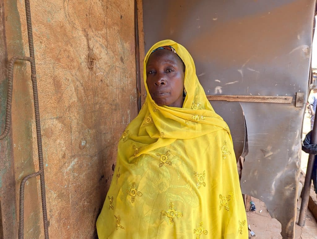 A woman in a vibrant yellow garment stands against a rustic wall, gazing thoughtfully at the camera.