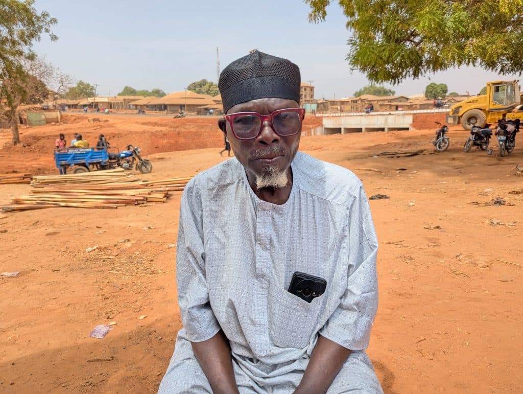 Man in a light outfit and red glasses sits outside, with a dirt road, quad bike, and motorcycles in the background.