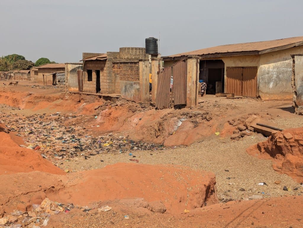 Rural scene with weathered buildings and a dry, litter-filled ditch under a clear sky.