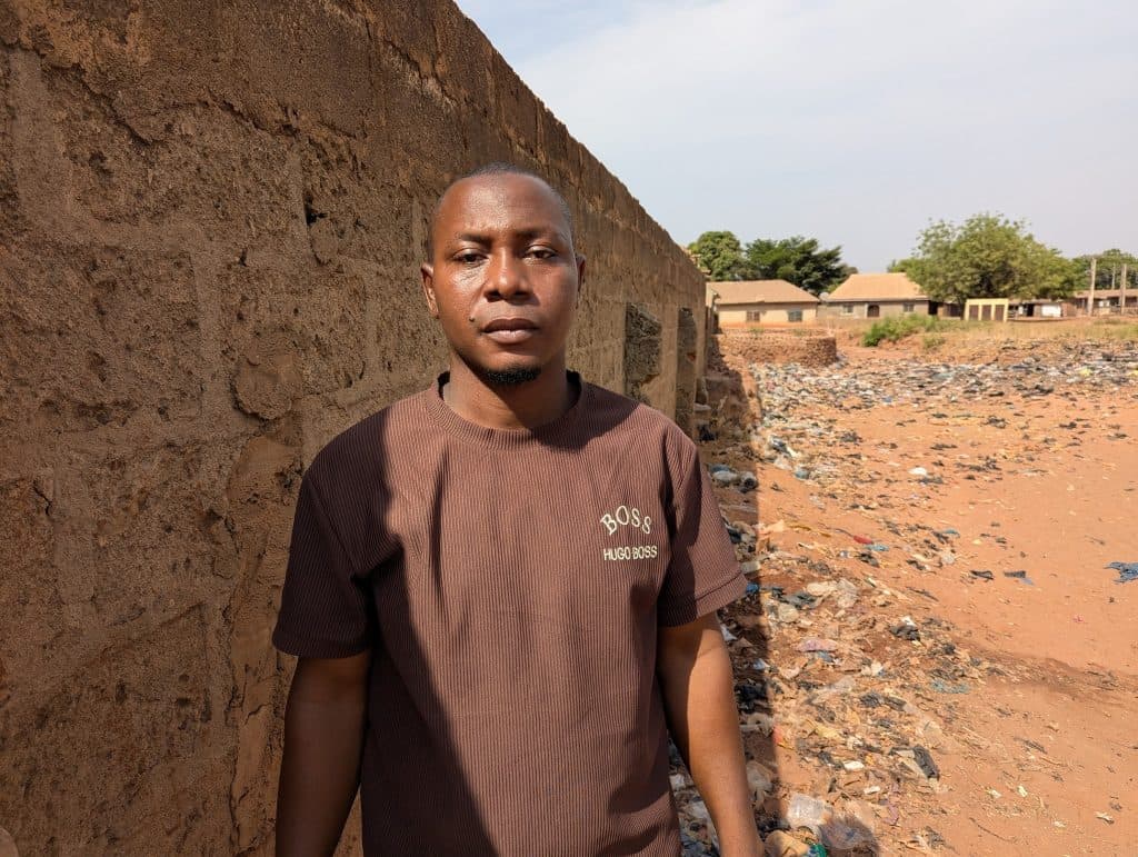 Man in a brown shirt stands next to a stone wall, with a littered ground and buildings in the background under a clear sky.