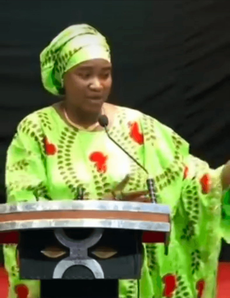 A person in a bright green patterned outfit speaks passionately at a podium with a microphone, gesturing with their hand.