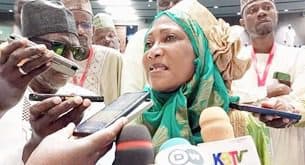 A woman in traditional attire speaks to a group of journalists holding microphones and recording devices.