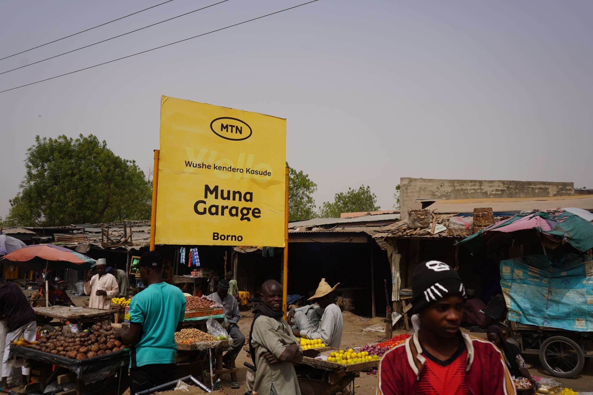 Outdoor market with a yellow "Muna Garage" sign in Borno. Vendors sell produce under makeshift stalls. People are browsing the market.