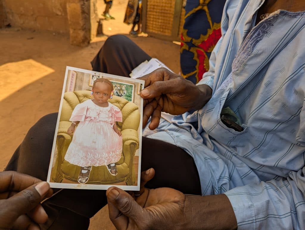 Hands holding a photo of a child in a pink dress, seated on a patterned chair. Background shows a small open area outside.