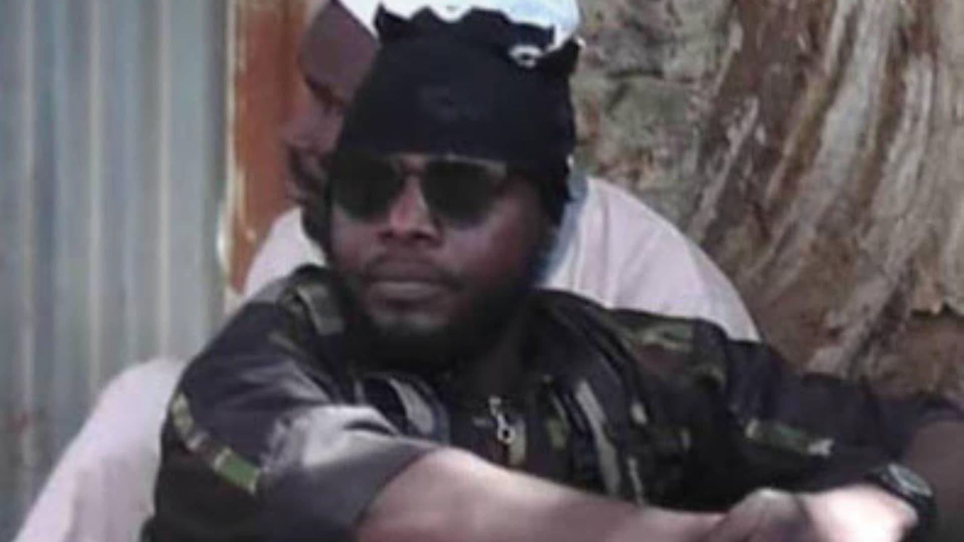 A person wearing sunglasses and a head covering sits outdoors with arms crossed, against a tree and a wall in the background.