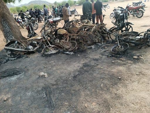 Burned motorcycle frames on the ground with people and intact bikes in the background.