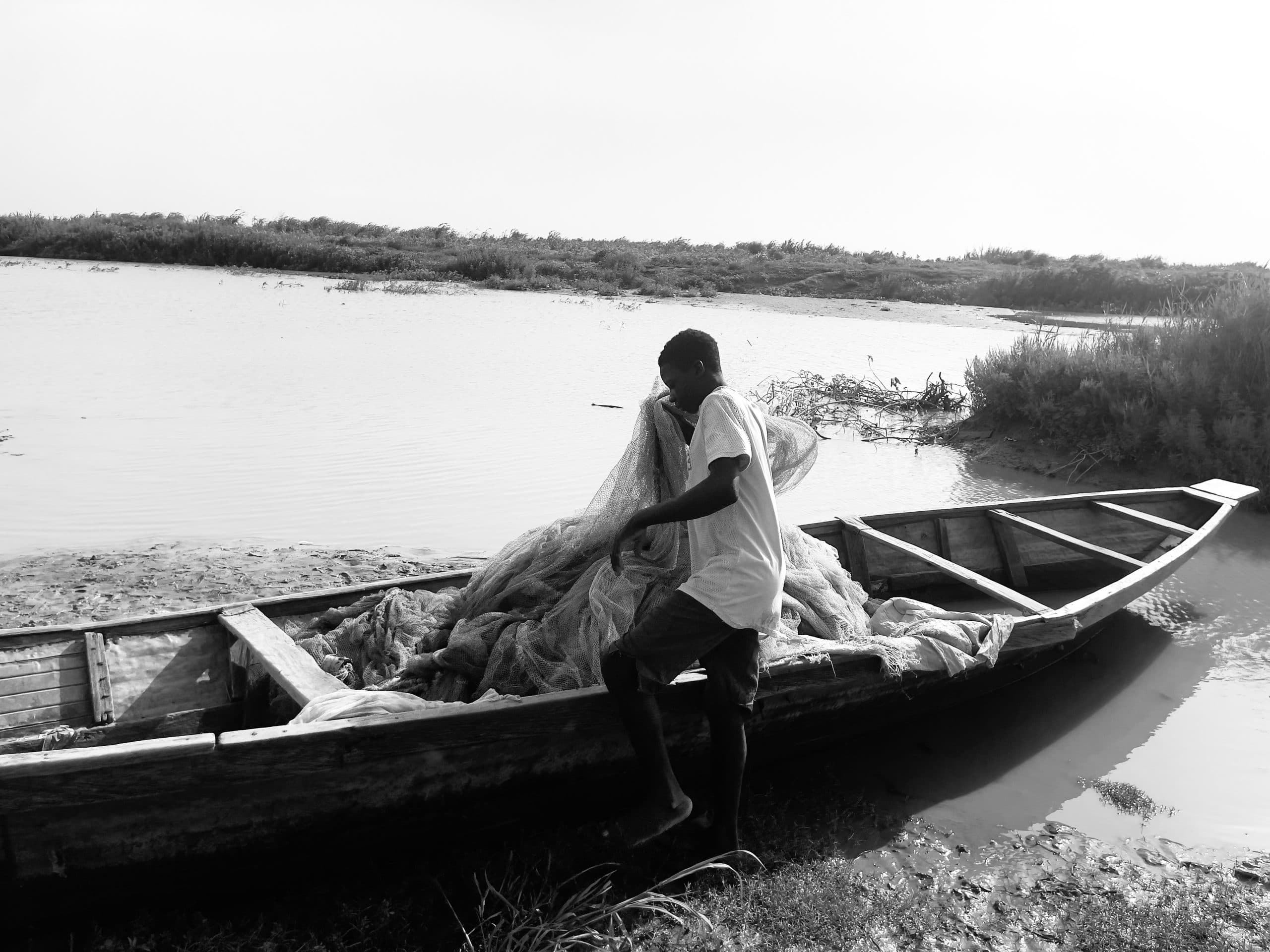 A person pulls in fishing nets on a small wooden boat by a riverside, surrounded by calm water and vegetation, in black and white.