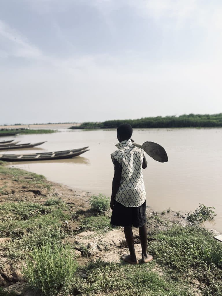 Person with a shovel looks at boats on a riverbank, surrounded by greenery and a cloudy sky.