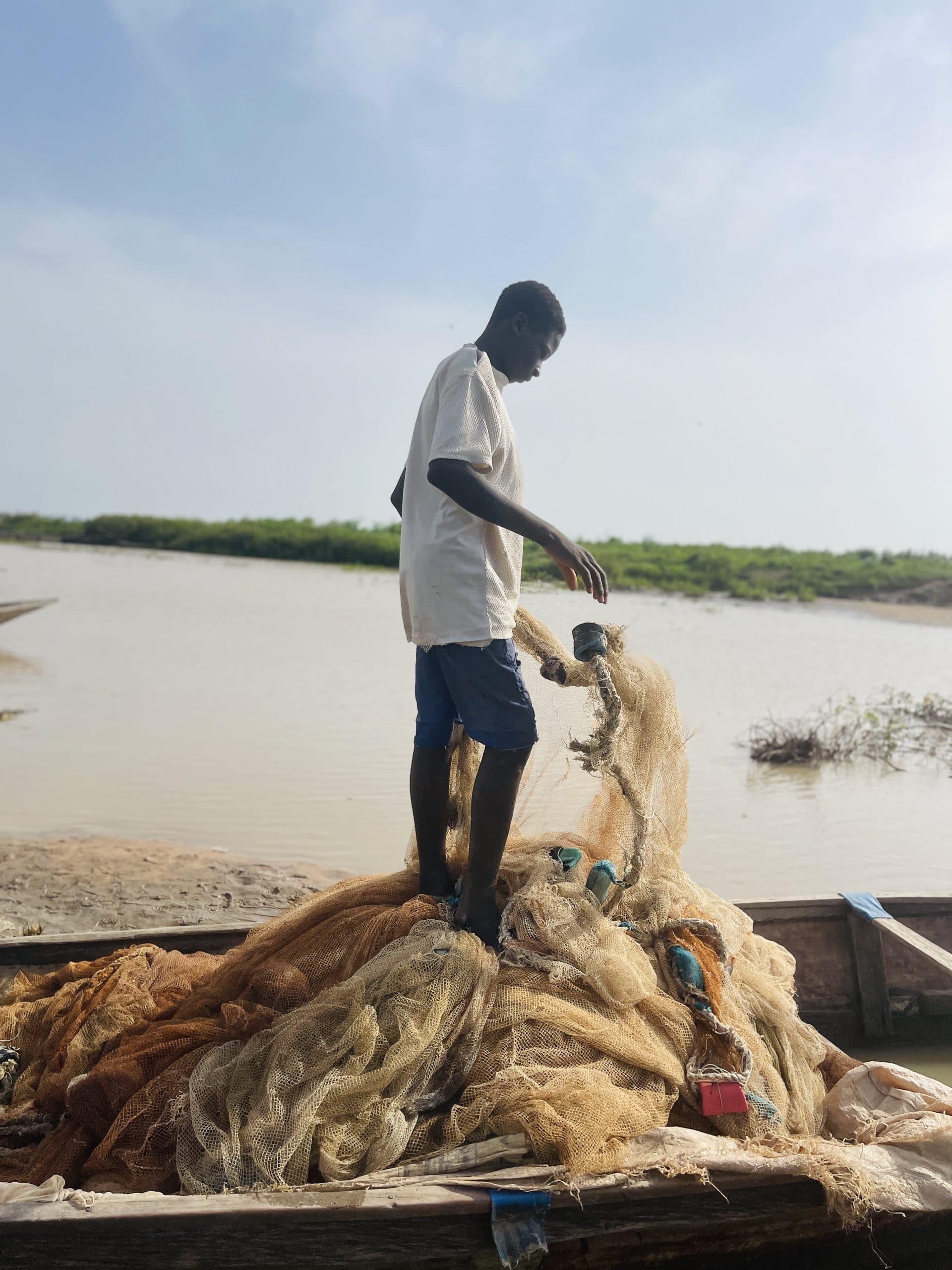 A person stands on a pile of fishing nets in a boat, by a calm river under a clear sky.