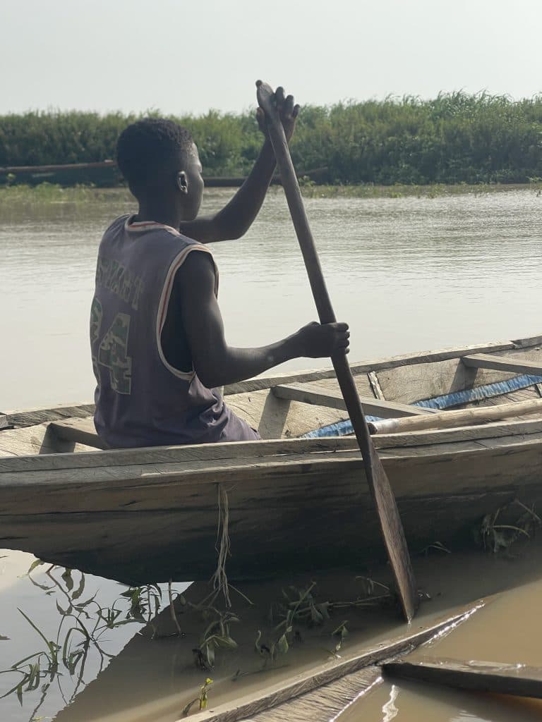 A person paddles a wooden boat on a calm river, surrounded by green vegetation under a clear sky.