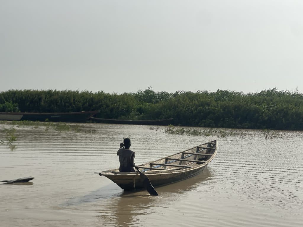 A person paddles a wooden boat on a calm river with lush greenery in the background.