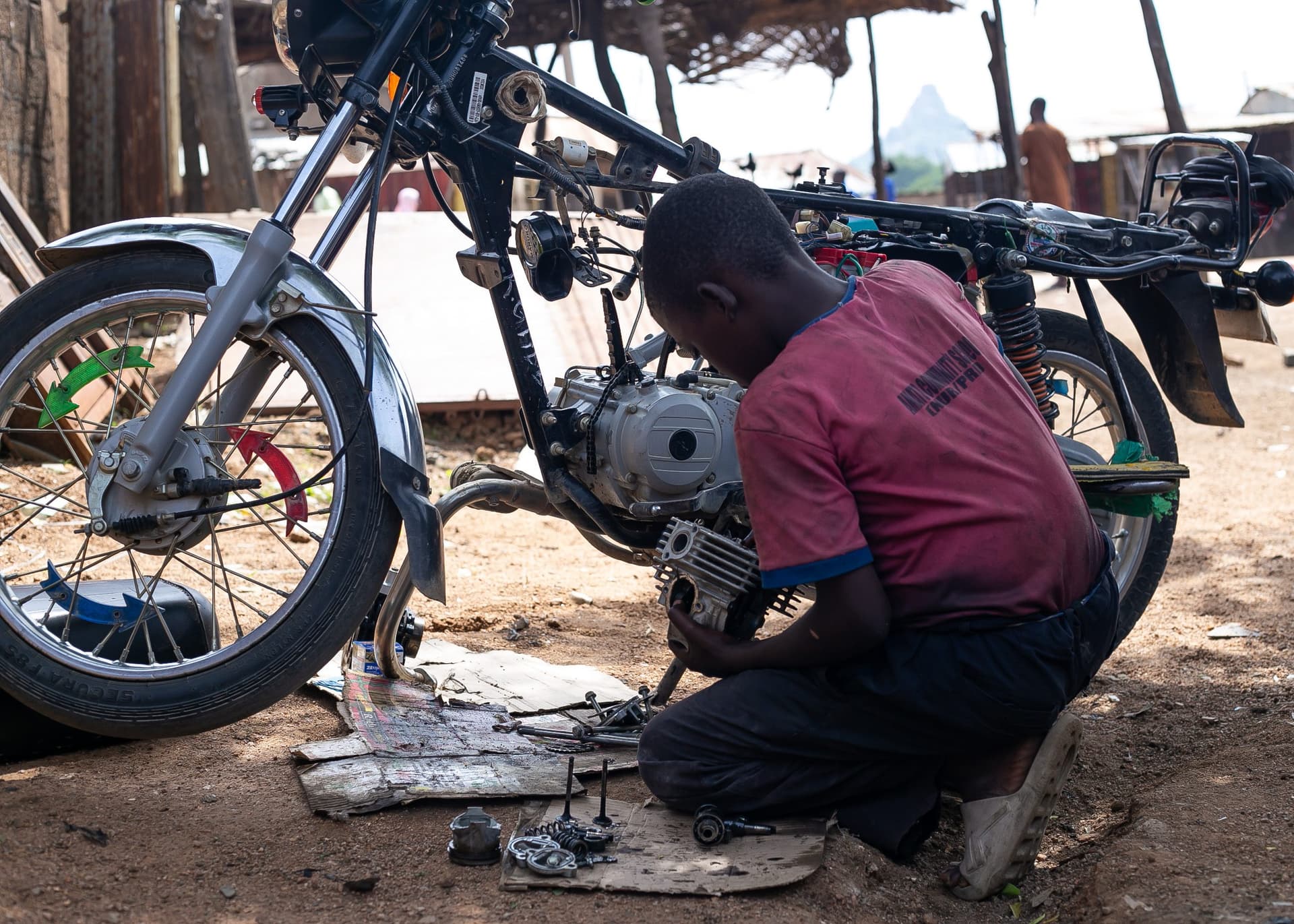 Young mechanic repairing a motorcycle outdoors, surrounded by tools and parts on the ground.