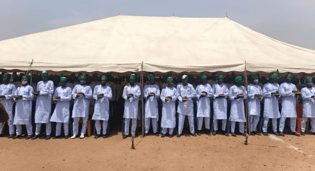A group of men in white outfits and green hats stand in formation under a large tent outdoors.