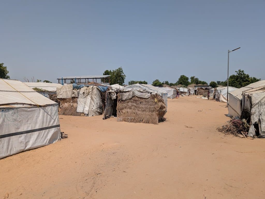 Rows of makeshift shelters with tarp and thatched roofs on sandy ground, under a clear blue sky. Sparse trees are visible in the background.