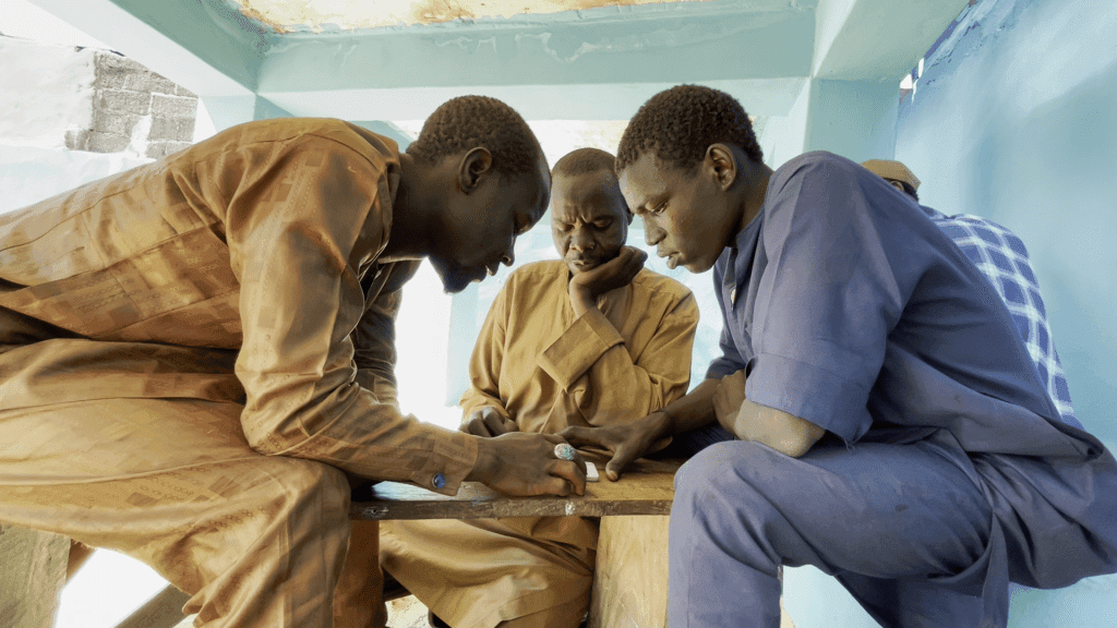 Three men in traditional clothing intensely focus on a small object on a wooden table in an enclosed space.