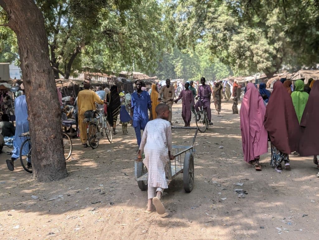 A bustling outdoor market scene with people walking, cycling, and a child pulling a cart under a canopy of trees.