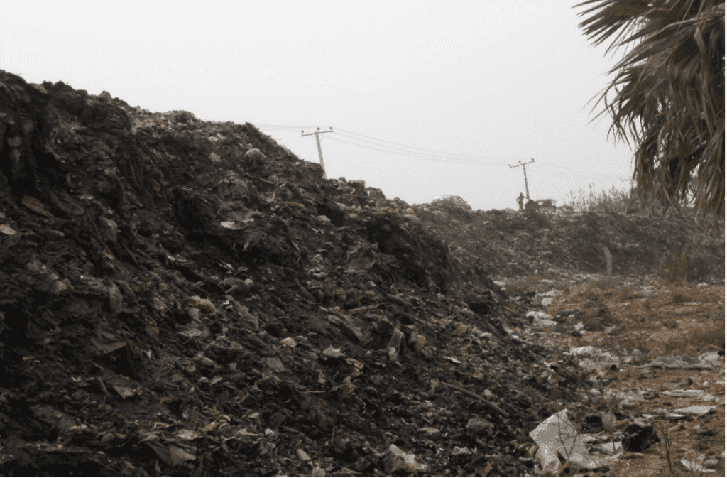 Large piles of garbage stretch along a dirt road, with power lines in the background under an overcast sky.