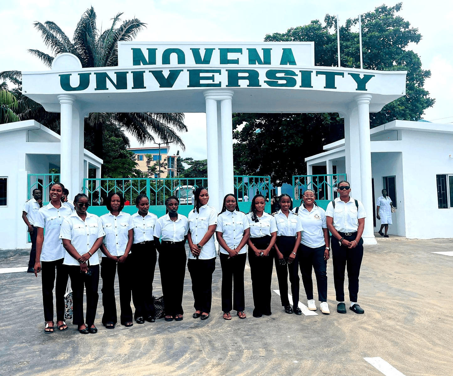 A group of people in uniform standing in front of Novena University entrance with a white gate and trees in the background.